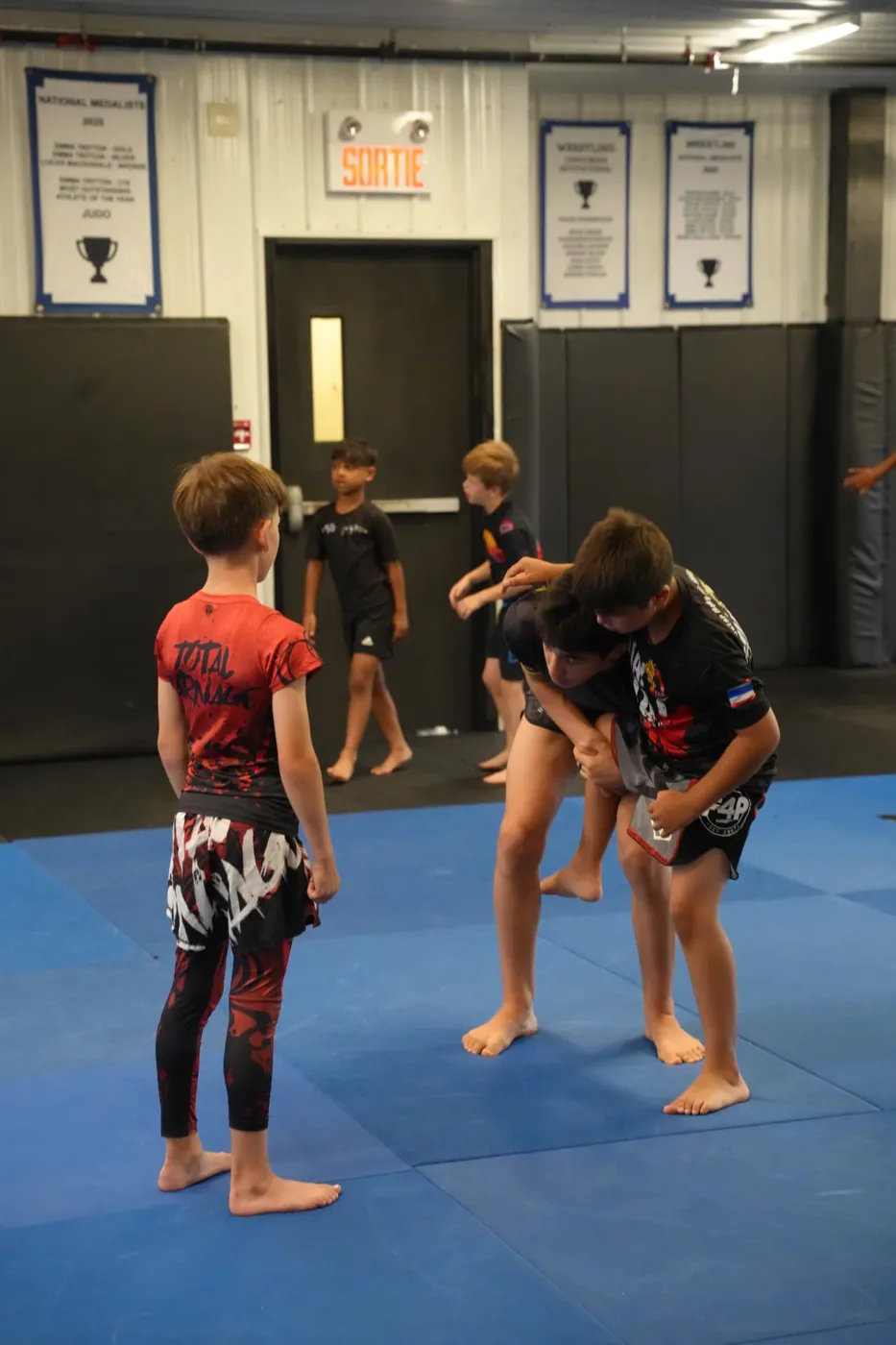 Kids working a body lock takedown while teammates train in the background